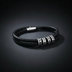 A black braided leather bracelet with silver-toned cylindrical beads featuring white lettering, displayed on a clean white background.