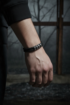 Male model forearm wearing double-wrap black leather bracelet with silver engraved beads in dark cinematic editorial photography